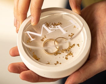 May include: White ring box with a clear lid, decorated with gold flakes. The lid has the inscription "M&H" in white lettering. Two gold wedding bands are inside the box.