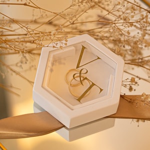 Könnte beinhalten: Weiße sechseckige Ringbox mit einem durchsichtigen Deckel, verziert mit den Initialen "V & T" in goldenen Buchstaben. Zwei goldene Ringe befinden sich in der Box.