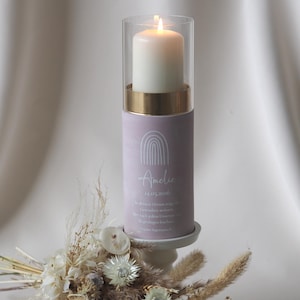 May include: A burning white candle in a clear glass cylinder sits on a lavender holder with gold trim. The holder is printed with the name "Amelie" and a date. A small bouquet of dried flowers is in front of the candle.