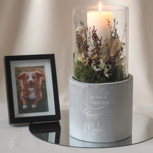 Op de afbeelding: Een brandende kaars in een glazen cilinder, omringd door gedroogde bloemen en groen, staat op een grijze betonnen basis. Links een ingelijste foto van een hond. De basis heeft tekst en een hand-vasthoudende afbeelding.