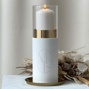 May include: A white candle holder with a lit candle inside a glass cylinder. The candle holder has a gold band and sits on a gold-colored tray. The base of the holder has an embossed design. Dried flowers are in the background.