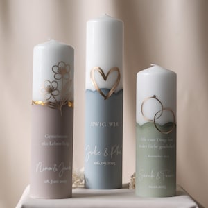 Puede incluir: Tres velas de pilar con diseños decorativos. Una vela tiene un corazón dorado, otra flores doradas y la tercera anillos de boda dorados. Cada vela tiene texto y fechas, con una combinación de colores blanco, azul y rosa.