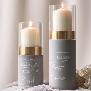 Peut inclure: Deux bougies piliers allumées dans des supports en verre et dorés. Les bases grises ont du texte et des illustrations blancs, dont un chat et une main tenant une autre main. Les bougies sont sur une surface claire.