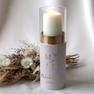 Puede incluir: Una vela blanca encendida dentro de un cilindro de vidrio transparente, sobre un soporte cilíndrico gris claro con detalles dorados. El soporte tiene letras doradas: "MD", un diseño floral y "Michaela & Denise 21/07/2020".