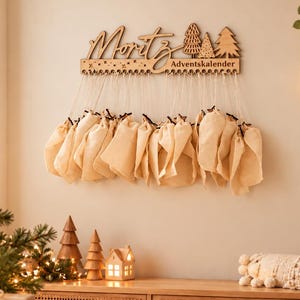 May include: A wooden wall-mounted advent calendar featuring the name Moritz in script, three decorative tree cutouts, and the text Adventskalender. Twenty-four small, light-colored fabric pouches are suspended from the wooden base by white strings.
