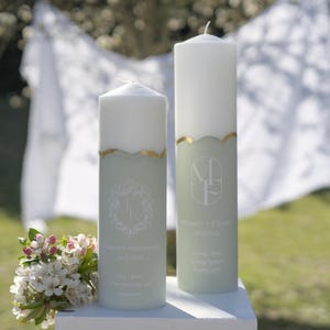 May include: Two white wedding unity candles with gold accents and personalized text. The left candle has a floral design and the text "Anna & Anthony 8.20.2023" and the right candle has the text "Vincent & Elena 8.20.2023" with a floral design.