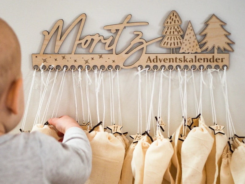 May include: A light-colored wooden wall-mounted advent calendar featuring the name Moritz in script, three stylized tree cutouts, and the text Adventskalender. It includes twenty-four small beige fabric pouches hanging from white strings.