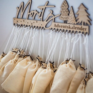 May include: A wooden wall-mounted advent calendar featuring the name Moritz and the text Adventskalender. The piece includes laser-cut tree and star motifs, with twenty-four white strings hanging down, each attached to a small beige fabric pouch.