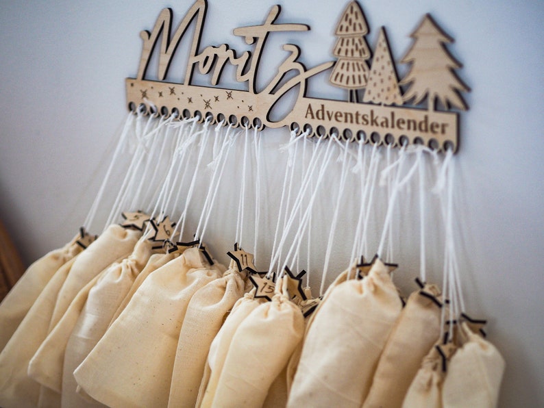 May include: A wooden wall-mounted advent calendar featuring the name Moritz and the text Adventskalender. The piece includes laser-cut tree and star motifs, with twenty-four white strings hanging down, each attached to a small beige fabric pouch.