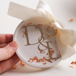 May include: White ceramic ring dish with gold lettering that says "D&L" and gold leaf accents. A white satin ribbon with a gold ring is on top of the dish.