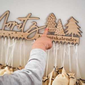 May include: A light-colored wooden wall-mounted advent calendar featuring the name Moritz in script, three stylized tree cutouts, and the text Adventskalender. Multiple small beige fabric bags hang from white strings attached to the wooden base.