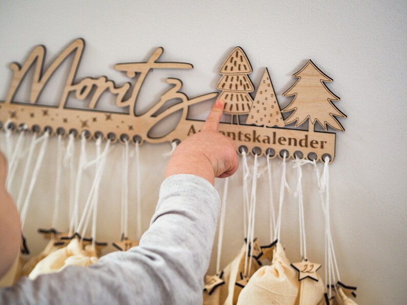 May include: A light-colored wooden wall-mounted advent calendar featuring the name Moritz in script, three stylized tree cutouts, and the text Adventskalender. Multiple small beige fabric bags hang from white strings attached to the wooden base.