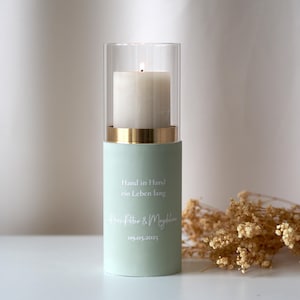 Puede incluir: Un portavelas verde claro con un cilindro de vidrio y un borde dorado. El portavelas tiene el texto "Hand in Hand ein Leben lang" y "Peter & Maylin 09.05.2023" impreso en él.