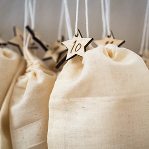 May include: A series of off-white fabric drawstring bags hanging from white strings. Each bag is attached to a small, light-colored wooden star ornament featuring a dark-engraved number, including the number 10 clearly visible on the front star.