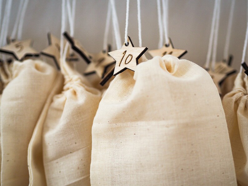 May include: A series of off-white fabric drawstring bags hanging from white strings. Each bag is attached to a small, light-colored wooden star ornament featuring a dark-engraved number, including the number 10 clearly visible on the front star.