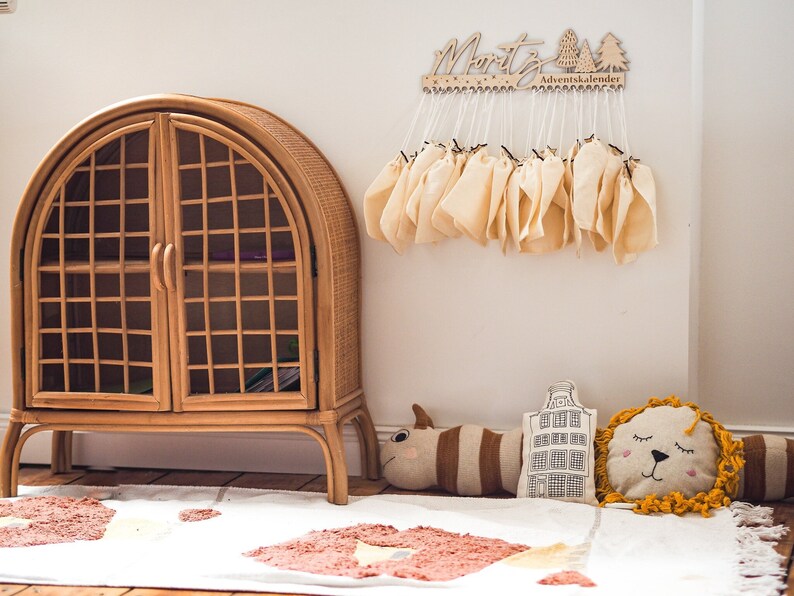 May include: A wooden wall-mounted advent calendar with the name Moritz and tree cutouts, featuring twenty-four small beige fabric drawstring bags hanging from white strings. Below, a rattan arched cabinet sits near a white rug with orange patterns.