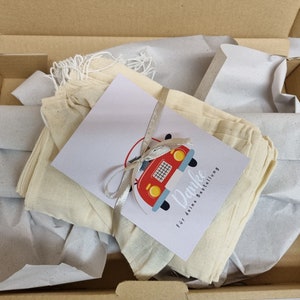 May include: A stack of off-white drawstring fabric pouches inside a cardboard box, wrapped with a white ribbon and a rectangular card featuring a red car illustration and the text Danke f&uuml;r deine Bestellung.