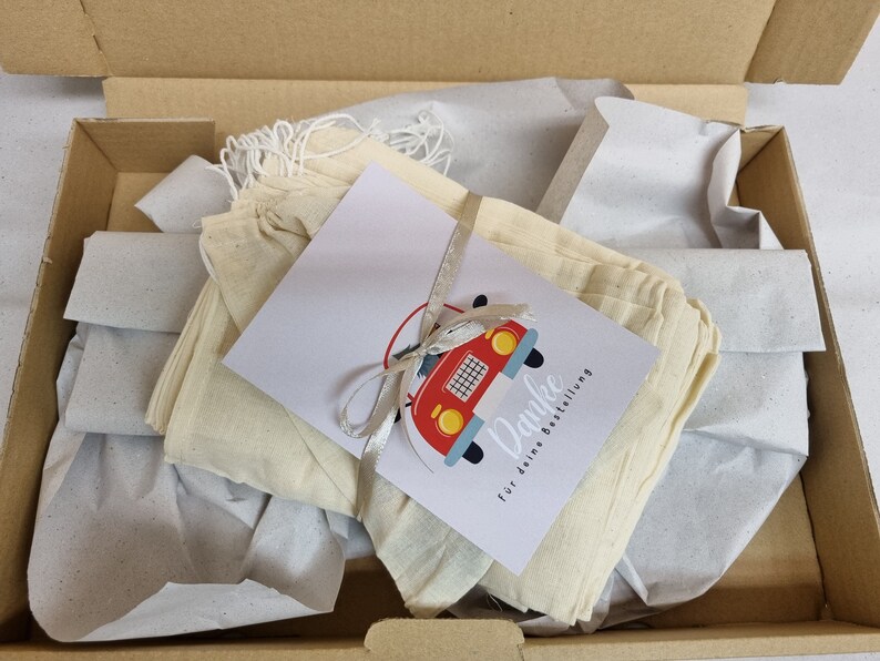 May include: A stack of off-white drawstring fabric pouches inside a cardboard box, wrapped with a white ribbon and a rectangular card featuring a red car illustration and the text Danke f&uuml;r deine Bestellung.