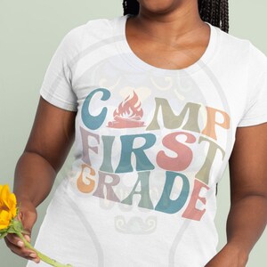 May include: White t-shirt with the text "Camp First Grade" in colourful retro font. The text includes a campfire graphic.