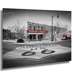 May include: A black and white photograph of a red truck parked in front of a building with a red sign that reads "Winslow, Arizona". The street is painted with a large Route 66 sign.