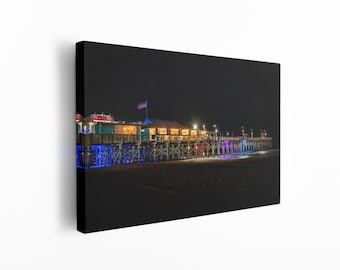 May include: A nighttime view of a pier with a restaurant and a bar on top. The pier is lit up with colorful lights, including blue, purple, and white. The pier is located on a beach with sand and water in the background.
