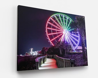 May include: A brightly lit Ferris wheel with red, green, and purple lights against a dark night sky. The Ferris wheel is located on a boardwalk with palm trees and a walkway in the foreground.