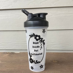 May include: A white shaker bottle with a black lid and a black floral design. The bottle has the text "Dead Inside But Caffinated" on it.