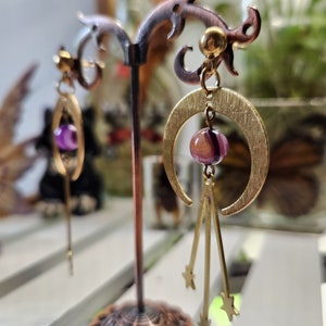 May include: A pair of gold crescent moon earrings with a purple agate stone and dangling stars. The earrings are displayed on a decorative stand.