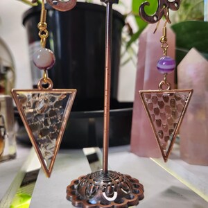 May include: A pair of gold-coloured earrings with a triangular pendant. The pendant has a textured, patterned design. Each earring features a small, round, purple and white bead. The earrings are displayed on a decorative copper-coloured stand.
