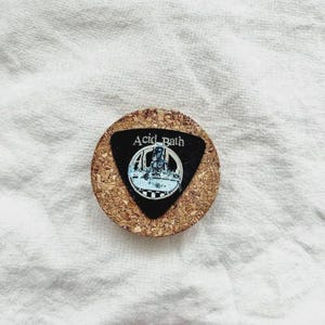 Pode incluir: Uma base de cortiça redonda com uma inserção triangular preta com o texto "Acid Bath" e um gráfico. A base é feita de cortiça natural e é adequada para bebidas.