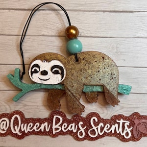 K&ouml;nnte beinhalten: Ein brauner und goldener Faultier, das an einem gr&uuml;nen Ast mit einer t&uuml;rkisfarbenen und goldenen Perlenkette h&auml;ngt. Das Faultier hat ein wei&szlig;es Gesicht mit schwarzen Augen und einem L&auml;cheln. Der Text "@QueenBea's Scents" befindet sich auf einem roten Glitzerhintergrund mit einer rosa Biene.