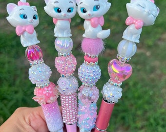 May include: Four pink and white jeweled pens with a white cat figurine on top. The cats are wearing pink bows. The pens are labeled #1, #2, #3, and #4.