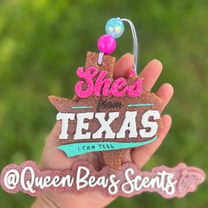 May include: A brown, Texas-shaped air freshener with the text "She's from TEXAS I CAN TELL" in pink, white, and teal. The air freshener has a string with pink and blue beads attached. The bottom of the air freshener has the text "@Queen Beas Scents".