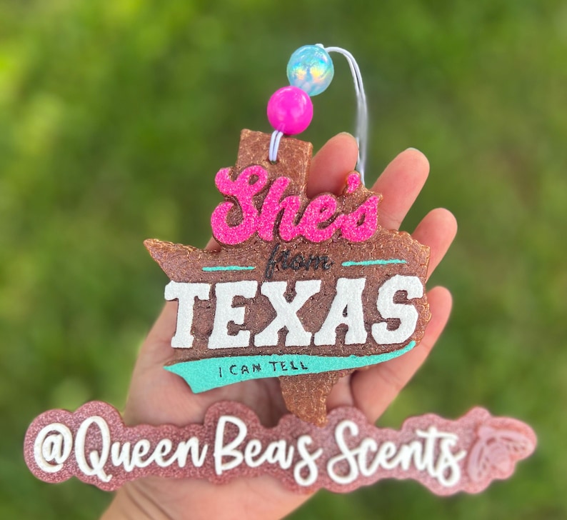 May include: A brown, Texas-shaped air freshener with the text "She's from TEXAS I CAN TELL" in pink, white, and teal. The air freshener has a string with pink and blue beads attached. The bottom of the air freshener has the text "@Queen Beas Scents".
