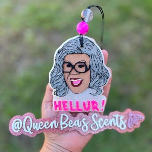 May include: A glittery air freshener featuring a cartoon image of a woman with glasses and gray hair. The air freshener has the text "HELLUR!" and "@Queen Beas Scents". It is attached to a black string with pink and silver beads.