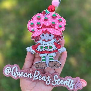 May include: A glittery Strawberry Shortcake character air freshener with a pink strawberry hat and red hair. The air freshener has a white dress with red and green accents. The text "@Queen Beas Scents" is below the character.