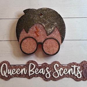 May include: A black and brown Harry Potter-themed wax melt with a lightning bolt scar and round glasses. The wax melt is on a white wooden background with the text "@Queen Bees Scents" in brown glitter.