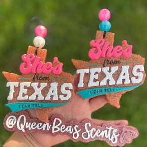 May include: Two Texas-shaped air fresheners with the text "She's from TEXAS I CAN TELL". The air fresheners are brown with white, pink, and teal accents. Each has a string of beads at the top. The bottom has the text "@Queen Beas Scents".