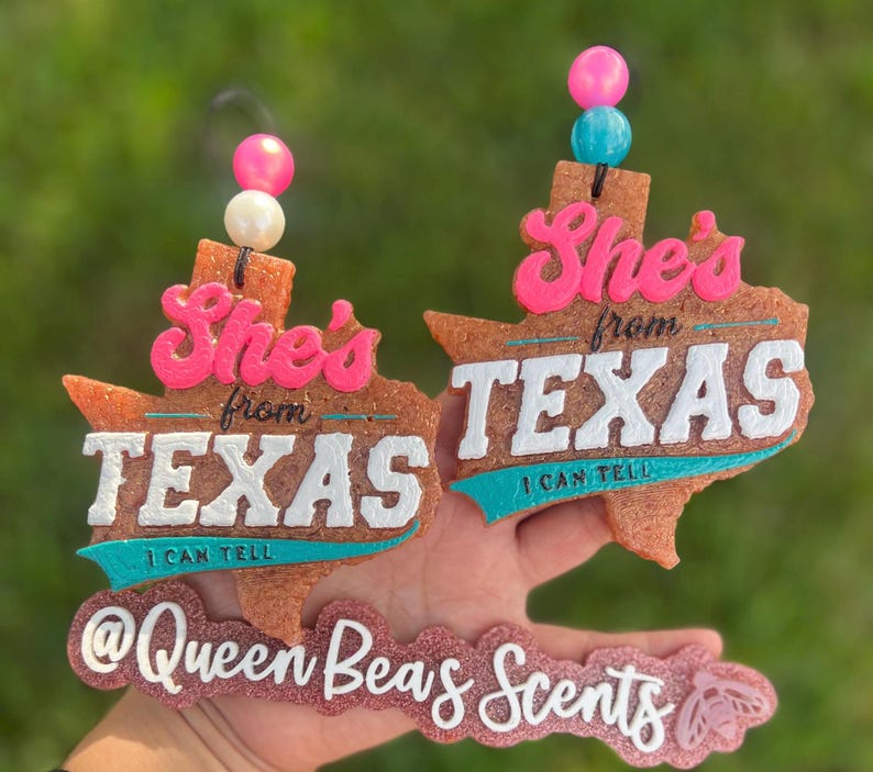 May include: Two Texas-shaped air fresheners with the text "She's from TEXAS I CAN TELL". The air fresheners are brown with white, pink, and teal accents. Each has a string of beads at the top. The bottom has the text "@Queen Beas Scents".