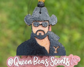 May include: Black and tan air freshener in the shape of a cowboy with sunglasses and a beard. The air freshener has the text "@Queen Beas Scents" in white on a pink glitter background. A black string and decorative beads are attached.