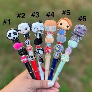 May include: A collection of six decorative pens, each featuring a different horror-themed character figurine at the top. The pens have various colors, including black, red, and blue, with decorative beads and accents.