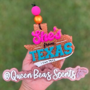 May include: A brown, Texas-shaped air freshener with the words "She's from TEXAS I CAN TELL" in pink and teal. The air freshener has a pink and orange bead accent and the text "@Queen Beas Scents" in pink.