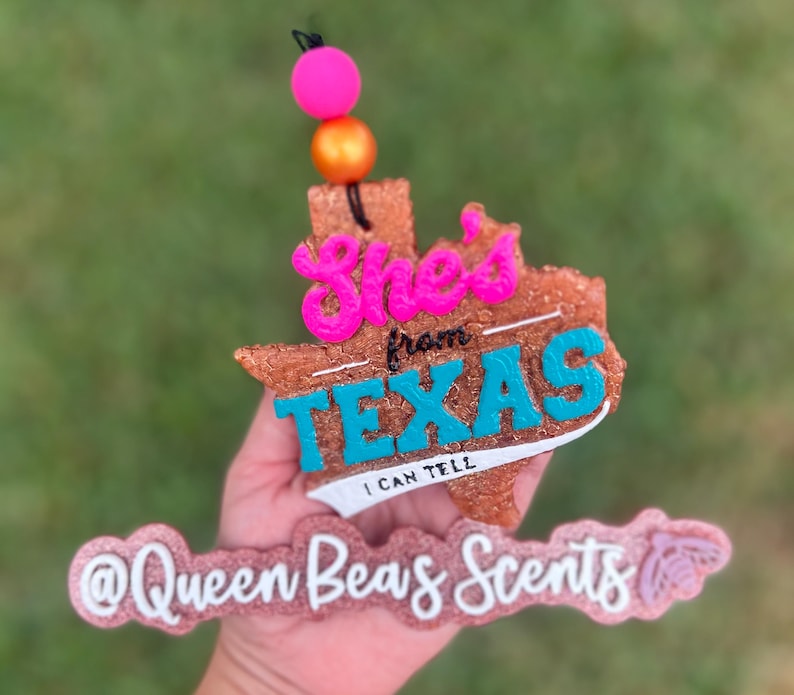 May include: A brown, Texas-shaped air freshener with the words "She's from TEXAS I CAN TELL" in pink and teal. The air freshener has a pink and orange bead accent and the text "@Queen Beas Scents" in pink.