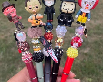 May include: Five different horror movie character themed pens with colorful beads and charms. The pens feature Freddy Krueger, Leatherface, Jigsaw, V for Vendetta, and Pennywise.