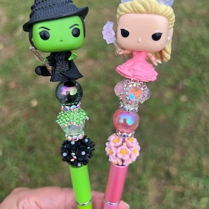 May include: Two green and pink pens with decorative tops featuring pop culture figures. The green pen has a black witch figure with a green face and a black hat. The pink pen has a blonde figure with a pink dress and a crown. Both pens have beads and flowers on the top.
