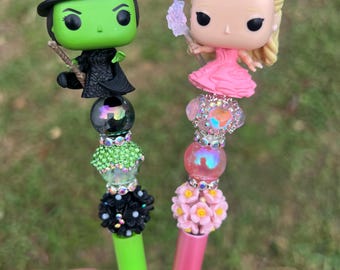 Op de afbeelding: Twee pennen, een groene en een roze, met decoratieve toppen met popcultuurfiguren. De groene pen heeft een zwart heksfiguur met een groen gezicht en een zwarte hoed. De roze pen heeft een blond figuur met een roze jurk en een kroon. Beide pennen hebben kralen en bloemen op de bovenkant.