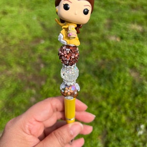 May include: A yellow pen with a decorative design. The pen features a Belle Funko Pop figure at the top, with brown, silver, and floral beads. The pen is held against a green background.