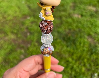 May include: A yellow pen with a decorative design. The pen features a Belle Funko Pop figure at the top, with brown, silver, and floral beads. The pen is held against a green background.