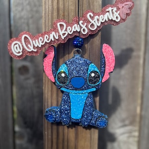 May include: Blue glitter Stitch character car air freshener with pink ears and a blue glitter bead hanging from a black string. The air freshener is hanging from a wooden fence post. The text "@Queen Bees Scents" is on a pink glitter banner.