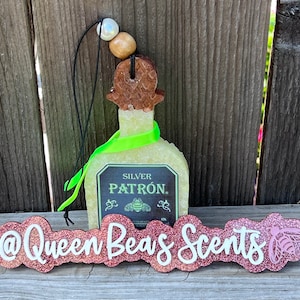 May include: A green and white Patron tequila bottle shaped car air freshener with a green ribbon and a wooden top. The air freshener is hanging from a black string with white and brown beads. The air freshener is in front of a wooden fence. The text "SILVER PATRÓN." is on the label of the bottle. The text "@Queen Bees Scents" is on a pink glitter sign with a bee graphic.